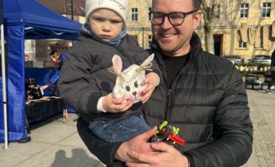 Uśmiechnięty mężczyzna z dzieckiem na rękach, trzyma znalezioną torebkę niespodziankę w kształcie zajączka. 
